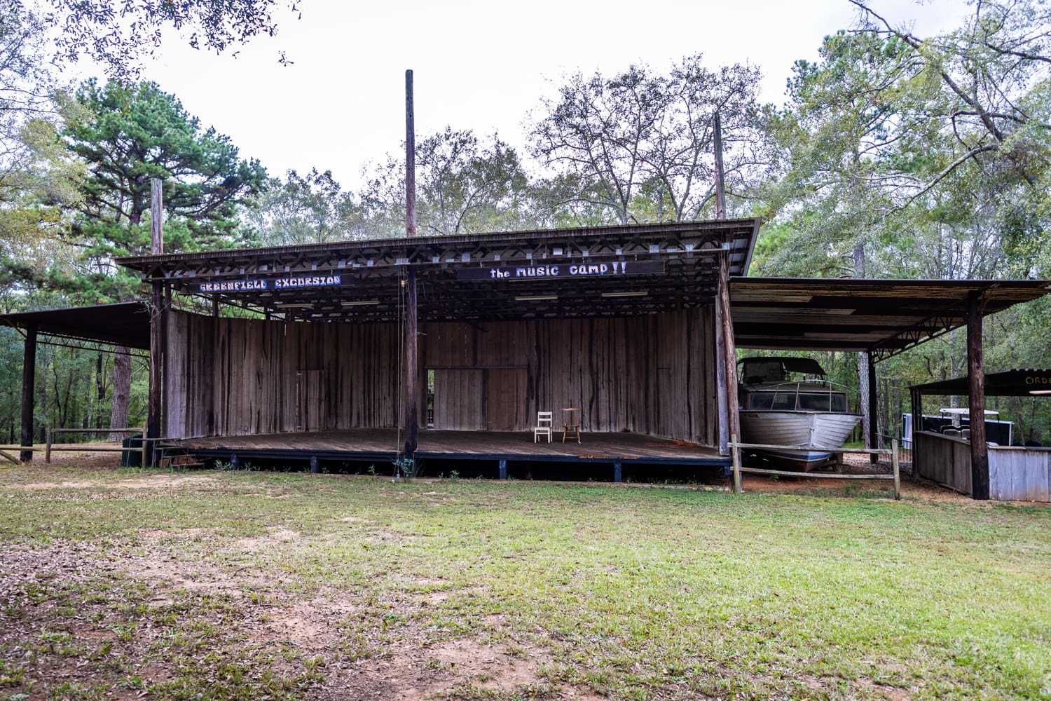 A view of the Greenfield Excursion stage, made from repurposed metal and wood materials. A sign reads "The Music Camp!!". Sitting beside the stage is an out-of-commission boat