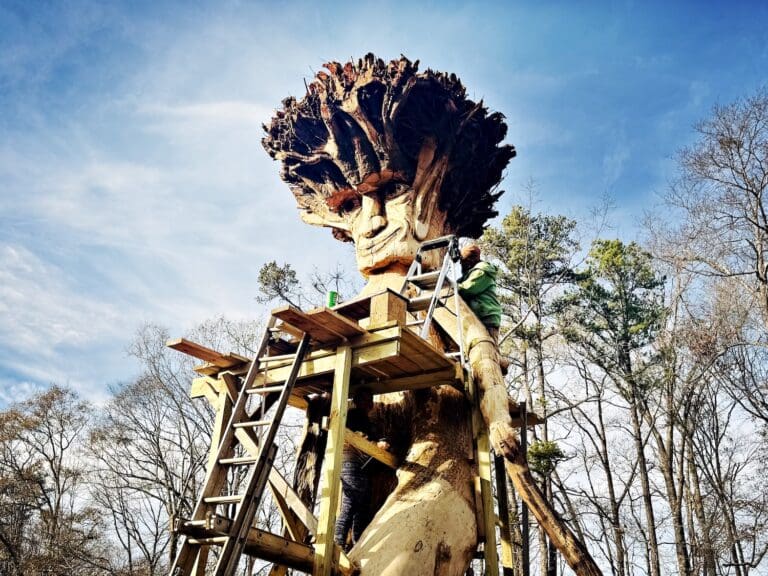 A large sculpture, still in progress, of a man carved in a tree. Artists are working on the top of the sculpture on ladders and scaffolding